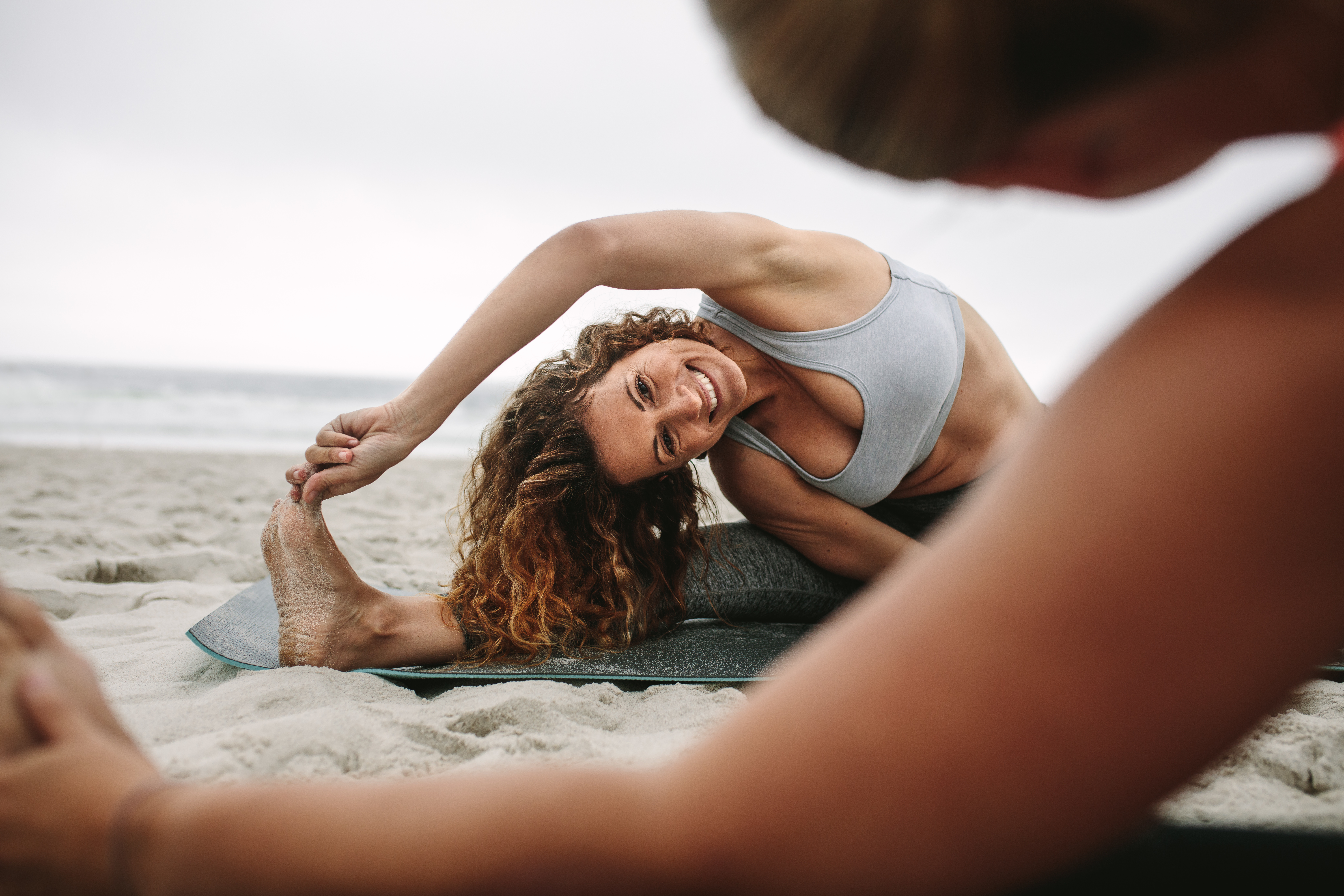 Fitness women sitting on yoga mats at the beach doing stretching workout. Women practicing stretching postures of yoga sitting near the sea.